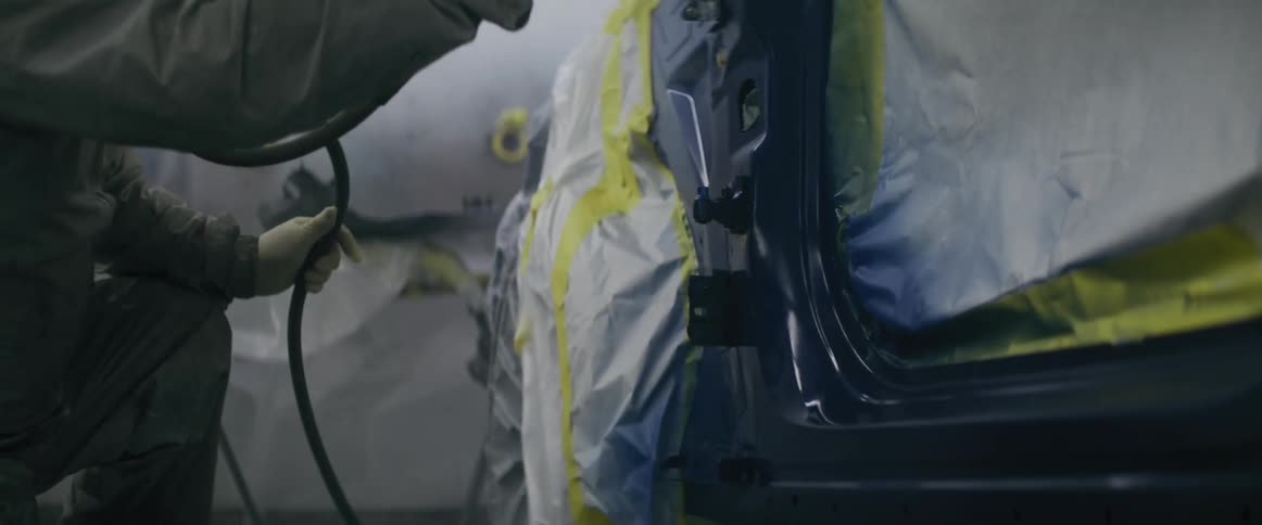 A worker in a grey jumpsuit uses a spray gun to paint a blue car door