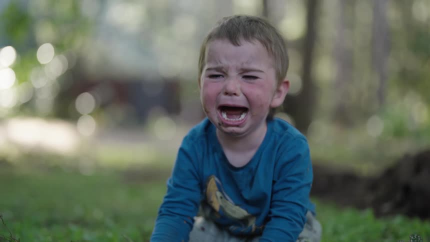 A male toddler screams and cries while sitting on grass outdoors