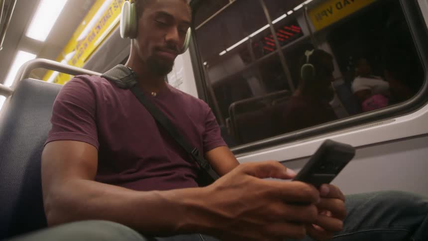 A young man sitting in a subway wearing headphones and holding a smart phone