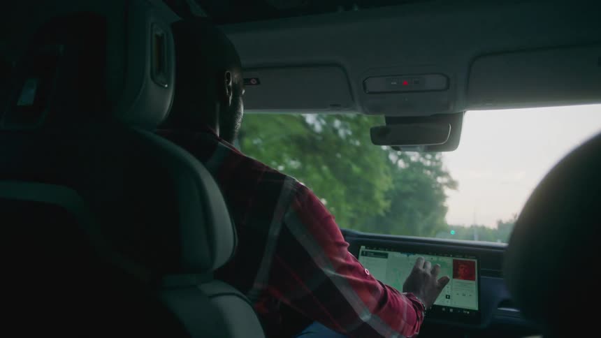 A man sitting on a car seat driving it and is fiddling with a multimedia screen