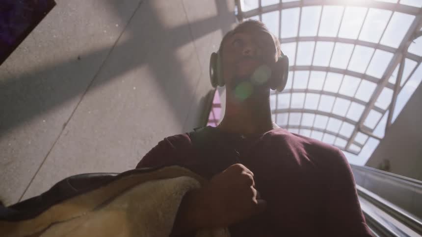 A young man with large headphones and holding a coat going down an escalator