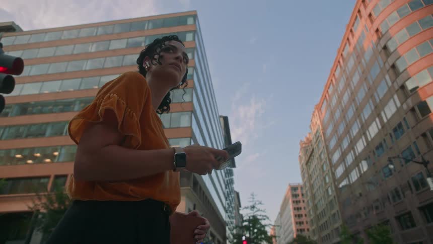 A woman in an orange shirt stands on the street using her phone and looking up at tall buildings