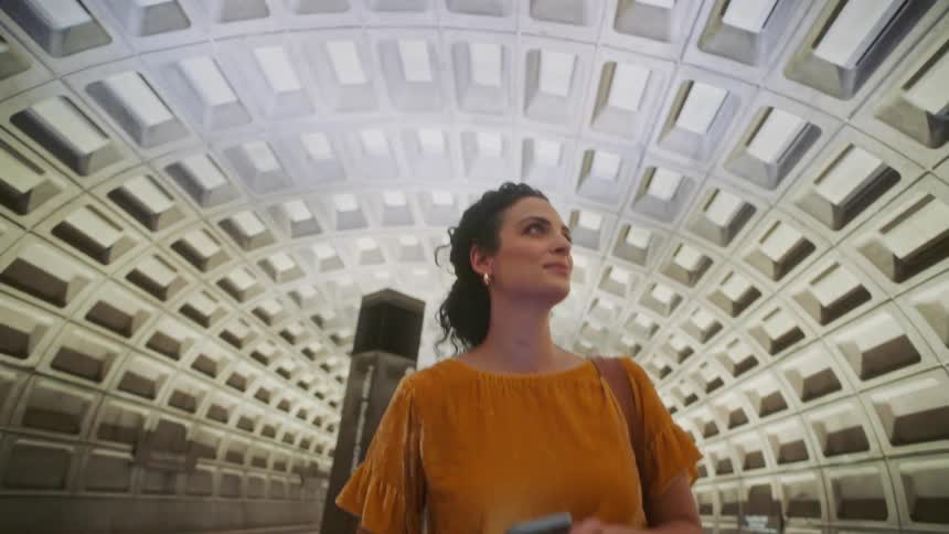 A woman in an orange blouse holding a cell phone in her hand is walking in a tunnelshaped indoor environment