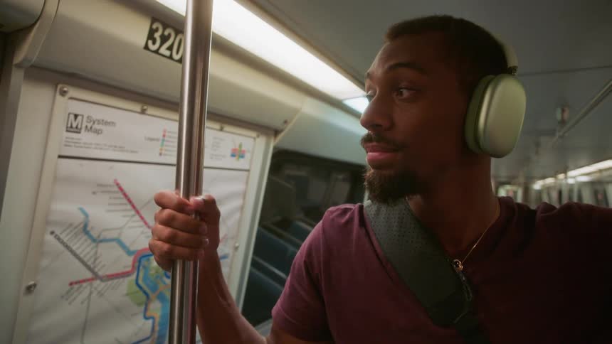 A man wearing headphones is holding onto a metal bar and he is inside a subway