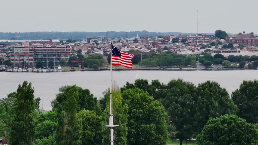 A large american flag waving proudly over a scenic waterfront city with a river buildings and trees in the background