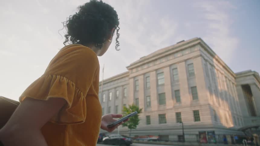 A woman wearing a yellow blouse holding a cell phone in her hand and looking ahead where there is a large white building