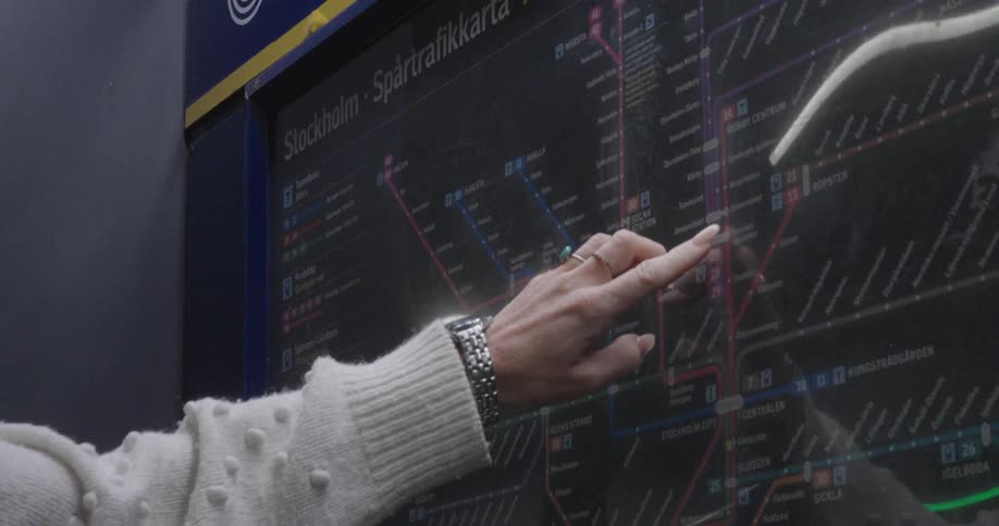 A woman stands in a rail terminal pointing at a map on a wall