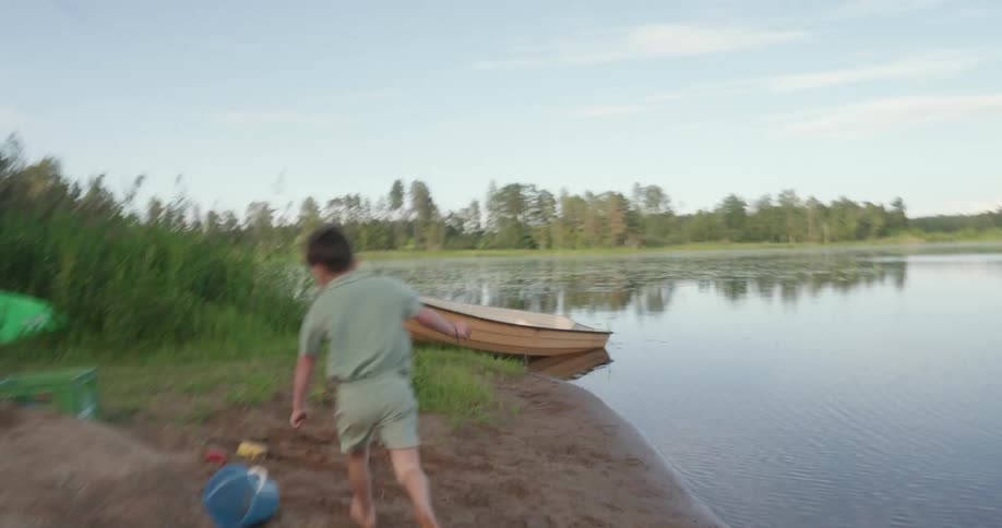 A young boy running all over a field of grass and mud next to a lake passing plastic toys and a canoe
