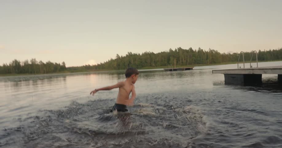 Boy splashes around in the water near a dock built across the water from the woods