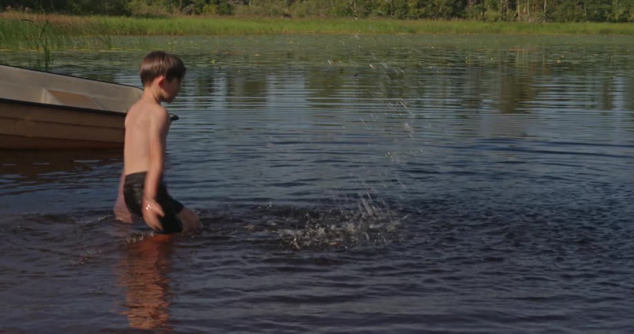 Boy standing in the shallow water near a boat throws water into the air