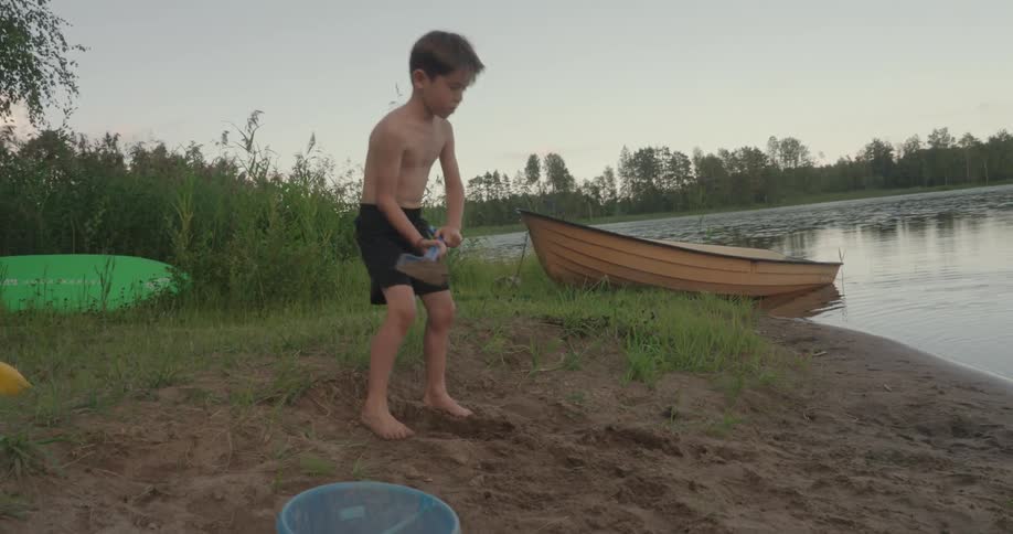 A young boy digging in the sand by a lakeside with a small blue shovel while a boat is moored nearby on the calm water surrounded by tall grass and trees