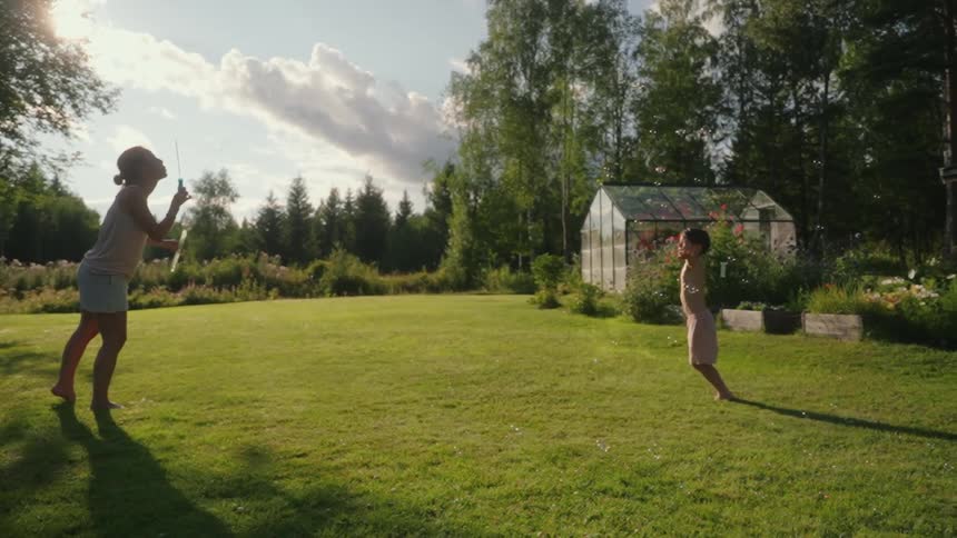 A woman standing in a large lawn blowing soap bubbles and a young boy trying to catch them