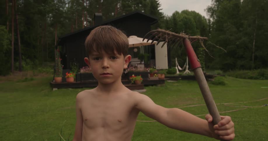 A boy standing in a grass yard looking serious and holding a rake