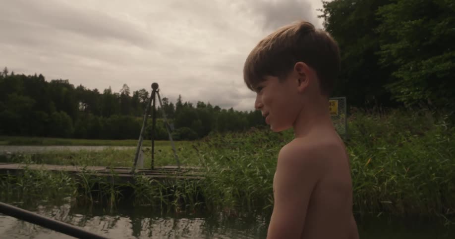 A shirtless boy fishing on a pond surrounded by vegetation