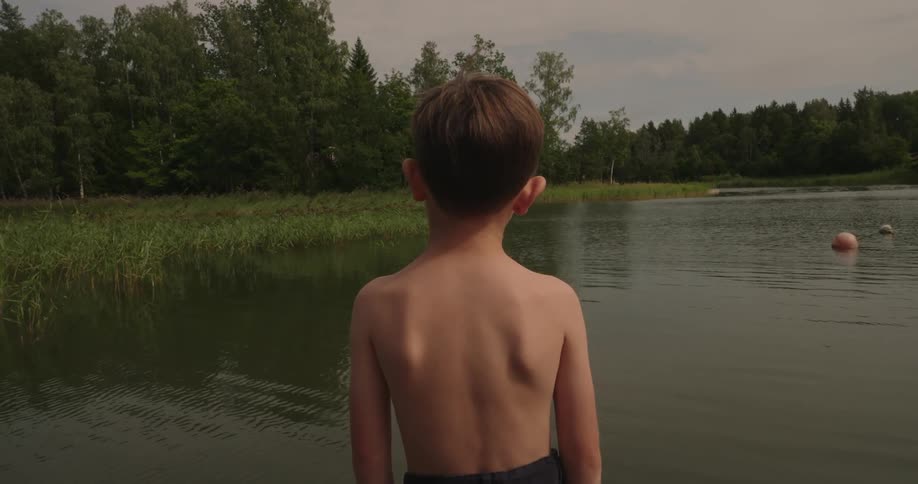 A young boy stands next to a small lake on a cloudy summer day