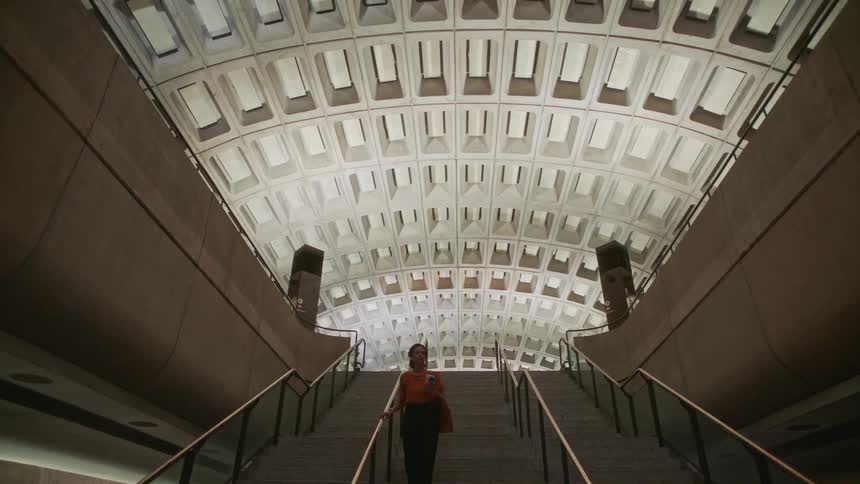 A woman going down a large flight of stairs inside a building while holding a plastic cup and a smart phone in the same hand