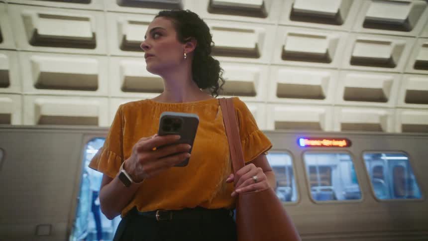 A woman in a yellow mustard top stands in a subway station holding a smartphone