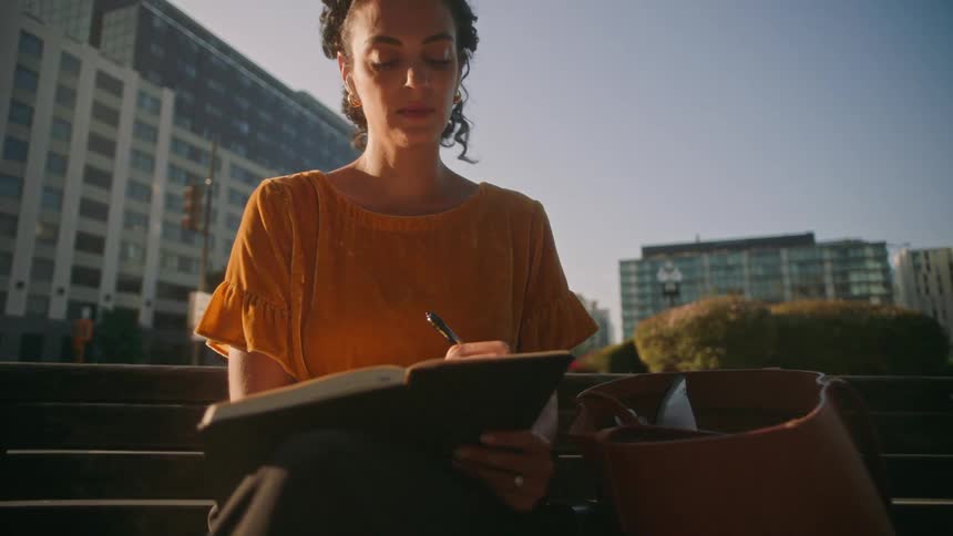 Woman sitting on a bench in a public square writing in a notebook