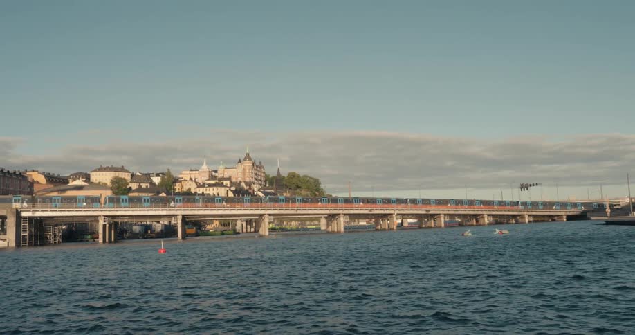 A train crosses a bridge over a calm river with a city with european buildings in the background