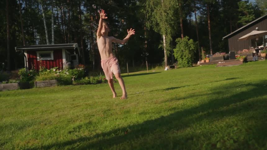 A boy running barefoot on a grassy lawn with a small red house and a greenhouse in the background