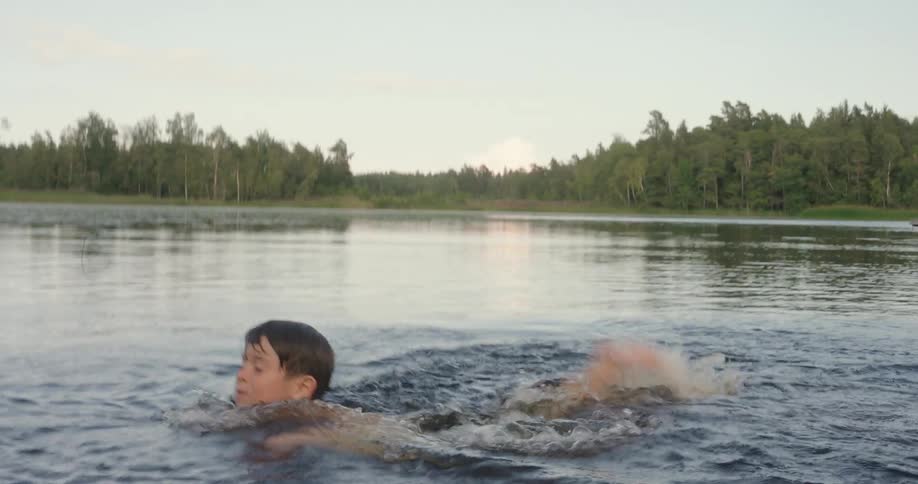 A young boy wearing dark swim trunks jumps from a wooden dock into a shallow lake where he swims briefly