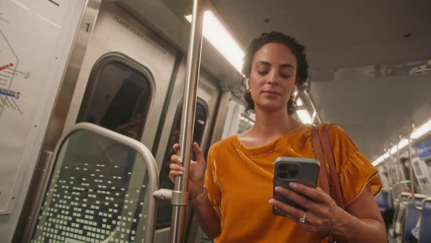 A woman in a mustard yellow top stands inside a train carriage holding onto a pole while listening to music on a smartphone