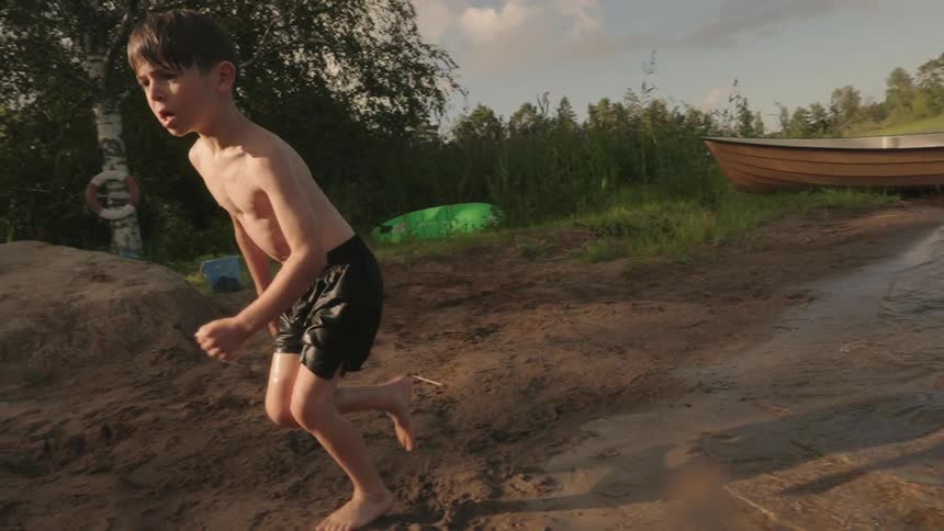 A young boy running out of a lake passing over mud to get to a lawn