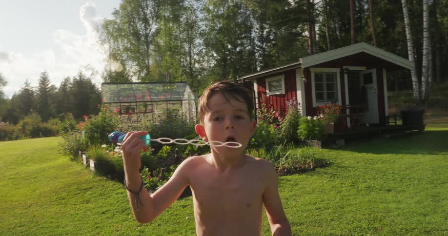 A shirtless little boy is in a grassy yard blowing an object that is making several soap bubbles