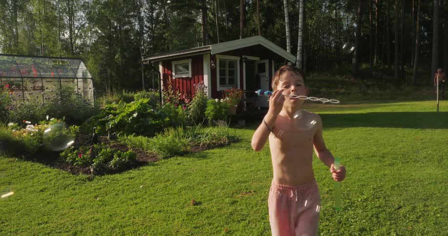 Boy in a backyard with a greenhouse in the background playing with soap bubbles