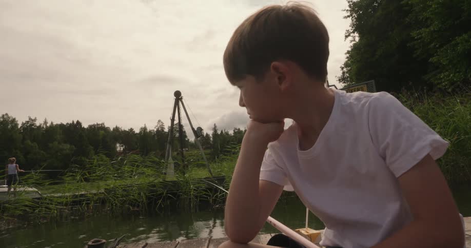 Bored looking boy sits on the dock and fishes in a pond near the woods on an overcast day