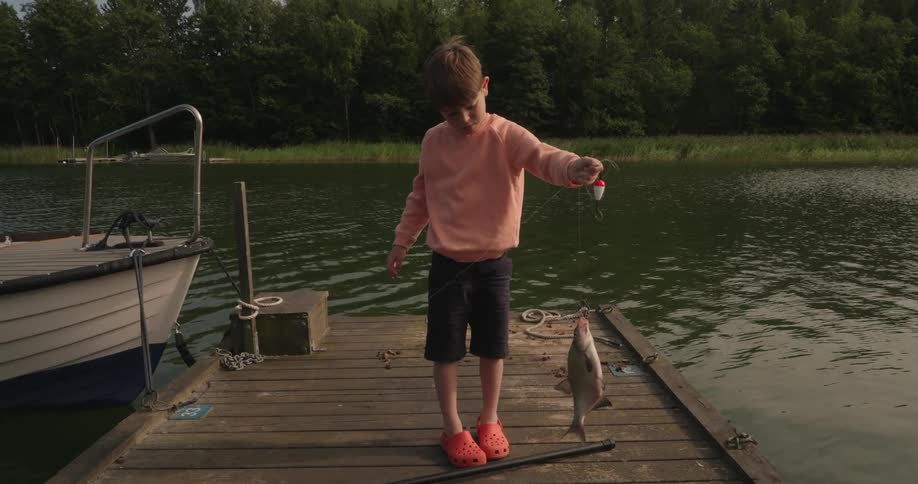 Boy stands on a dock across the river from the woods and holds a fish on a line
