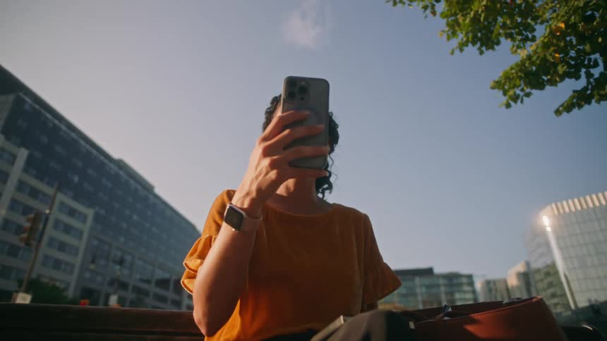 Woman with curly hair and orange t shirt sits on a bench using a smartphone with apartment buildings in the background