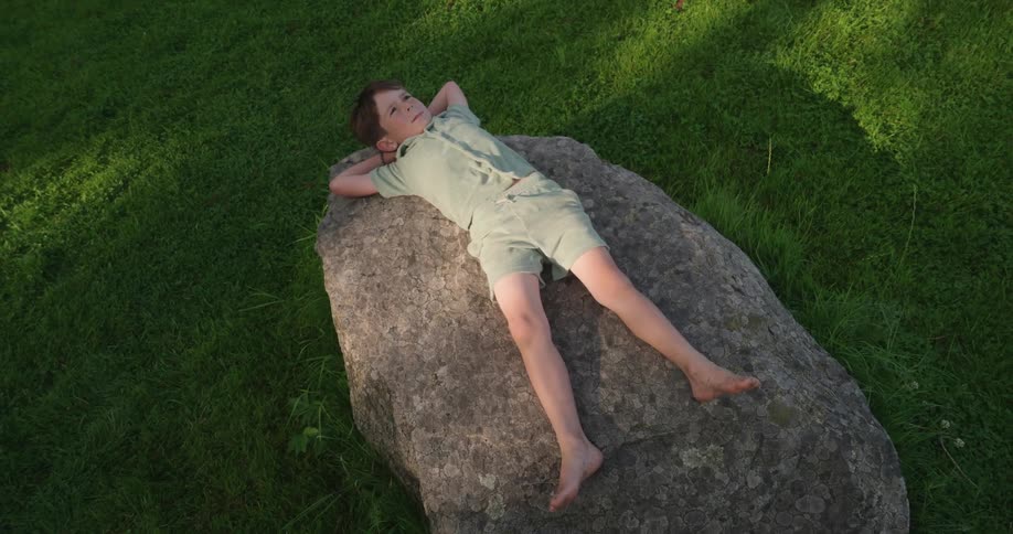 A young boy in shorts and a shirt lies on a rock and looks up into the sky