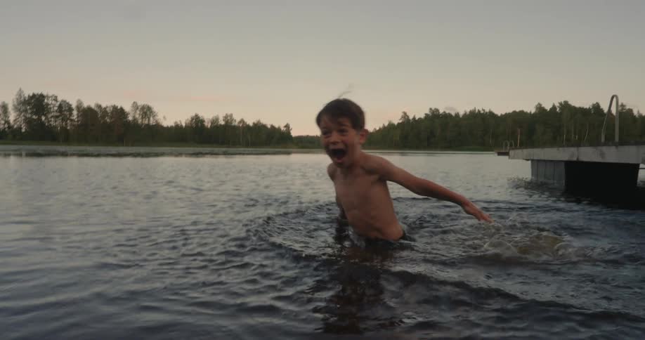 A young boy jumps into a lake and screams and laughs from the shock of the water temperature