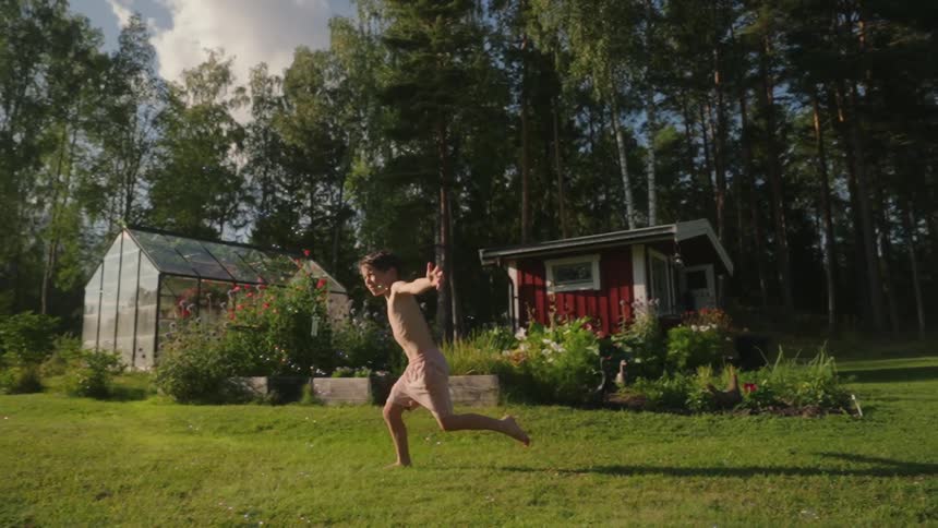 A boy running barefoot on a grassy lawn with a small red house and a greenhouse in the background
