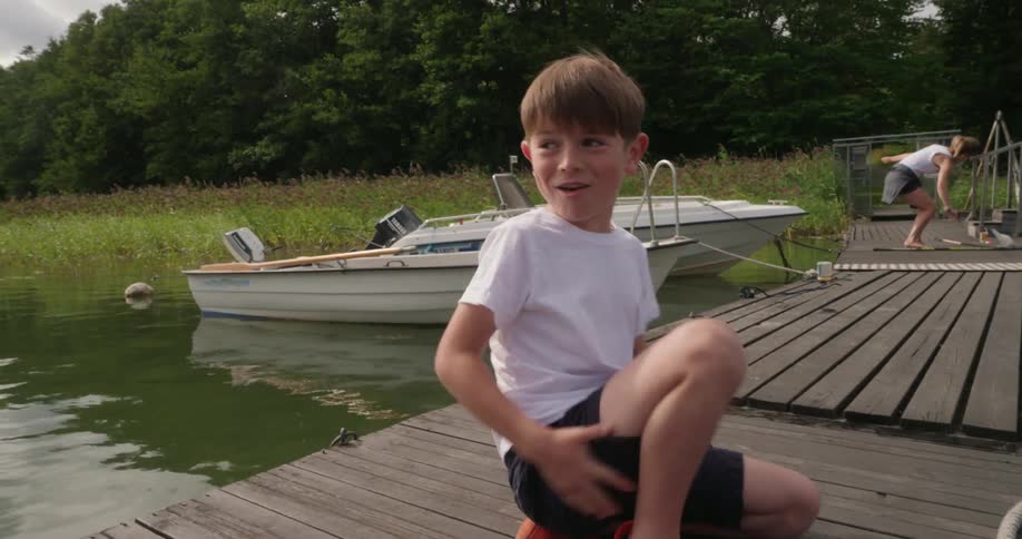 A young boy wearing a white shirt and navy shorts sitting on a wooden dock by the water with a boat moored nearby while another person is walking in the background