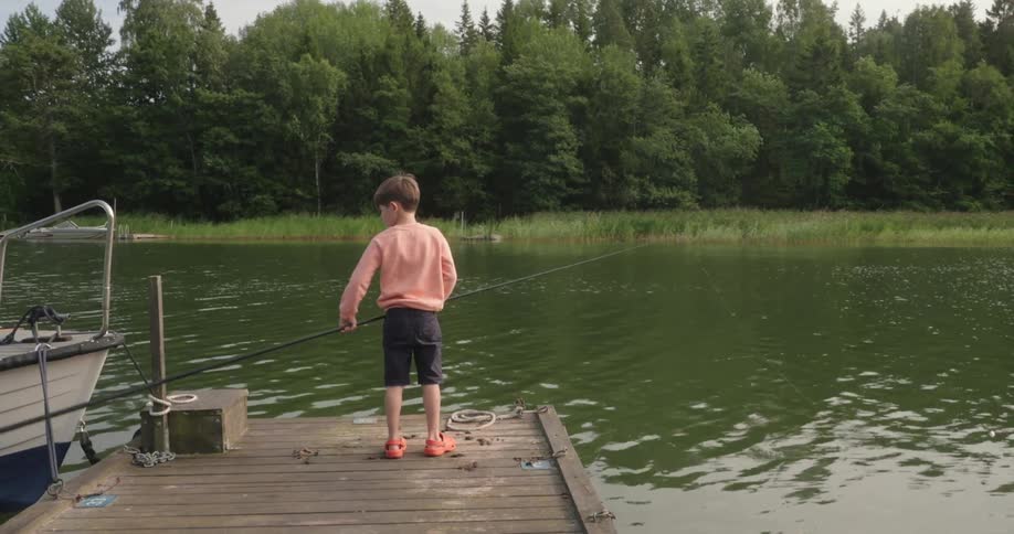 A little boy stands on the end of a wooden pier holding a fishing rod next to a boat