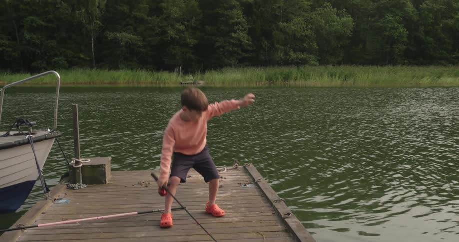 A boy stands on a wooden dock by a lake holding up a fishing rod
