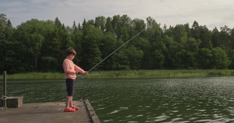 A young boy stands patiently on a wooden dock holding a fishing rod over a calm lake
