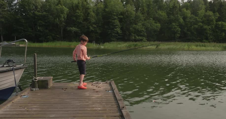 A young boy stands on a dock next to a moored boat as he casts a fishing line into a lake