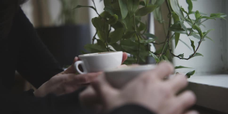 Two people sit indoors gripping their white coffee cups