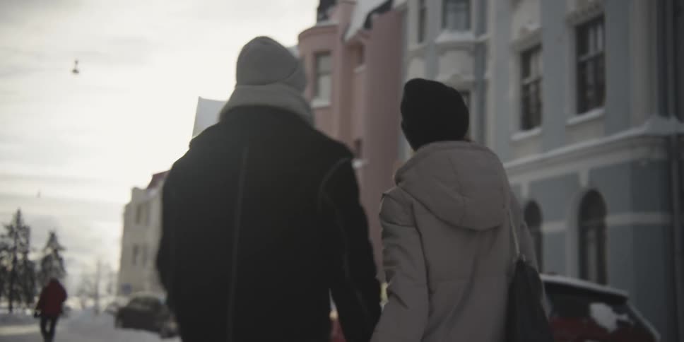 Back view of a man and a woman holding hands walking through the city on a snowy day