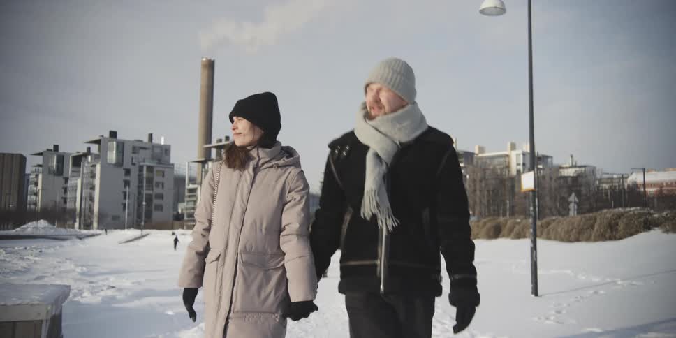 A couple walk hand in hand through a snowy landscape with a city a tall chimney in the background