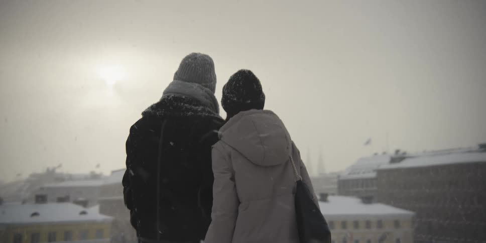A couple walk hand in hand through a snowy landscape with a city a tall chimney in the background