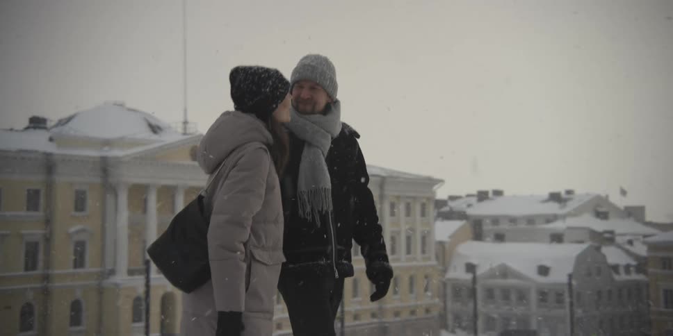 A man and a woman walk through falling snow in a city while holding hands