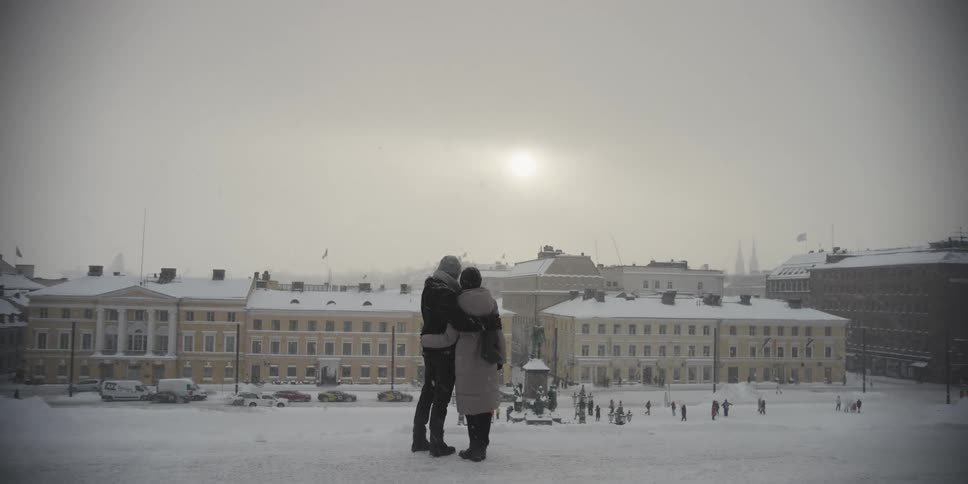 A couple stand close together in the foreground of a snowy cityscape overlooking a public square