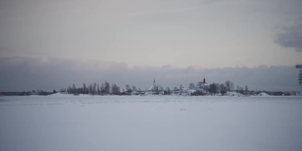 Landscape of a vast field covered in snow with houses and trees in the background