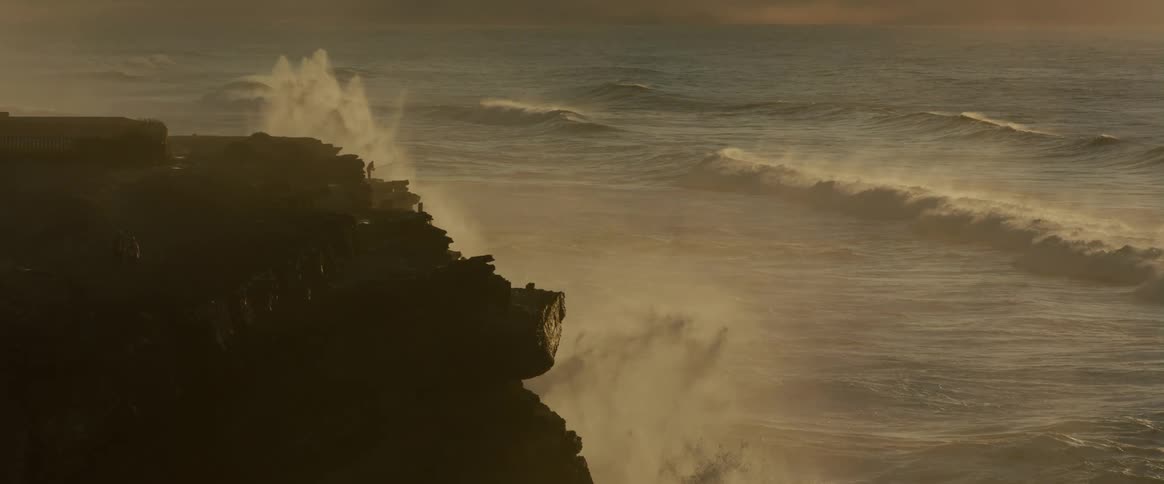 A person stands on a cliff while ocean waves crash against it in slow motion