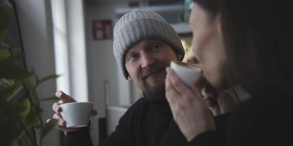 A man in a gray cap is talking to a woman who is next to him and they are both drinking something from a white mug