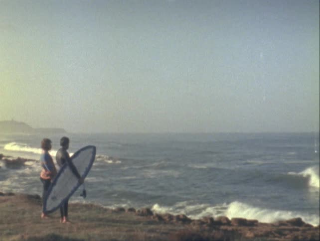 Man holding a surfboard next to a woman as they look out at the sea view